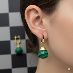 Puede incluir: Pendientes dorados con una piedra de malaquita verde en forma de lágrima. Los pendientes tienen un cierre de palanca y una tapa dorada. La piedra de malaquita presenta un patrón de rayas. Un pendiente está enfocado, con otro pendiente visible en el fondo.