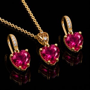Op de afbeelding: Een gouden sieradenset bestaande uit een ketting en twee oorbellen. Elk sieraad bevat een hartvormige, felroze edelsteen gezet in goud met kleine heldere steentjes op het oogje. De ketting is voorzien van een fijne gouden schakelketting.