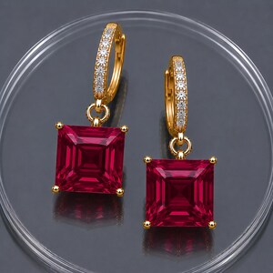 May include: A pair of gold hoop earrings featuring a row of small clear stones along the front and a square-cut deep red gemstone dangling from each hoop. The earrings are designed with a hinged closure and a polished gold metal finish.