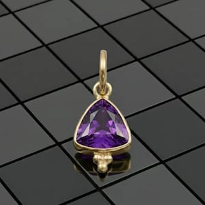 May include: A gold pendant featuring a triangular, faceted, deep purple gemstone. The pendant has a small loop at the top for a chain and a decorative cluster of three small spheres at the bottom. The setting is gold.