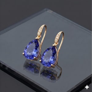 Könnte beinhalten: Ein Paar goldfarbene Ohrhänger mit großen, birnenförmigen, tiefblauen Edelsteinen. Die Ohrringe haben ein gebogenes Haken-Design mit kleinen, klaren Akzentsteinen. Die Ohrringe werden auf einer reflektierenden Oberfläche präsentiert.