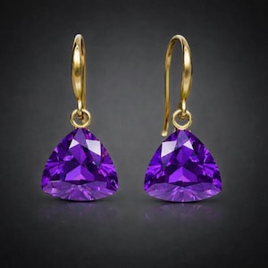 May include: A pair of gold-coloured drop earrings. Each earring features a large, triangular, faceted purple gemstone. The earrings have a simple hook design. The gemstones are set against a dark background, highlighting their vibrant colour.