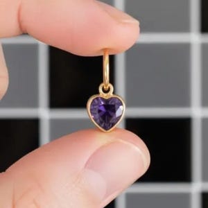 May include: A gold-coloured heart-shaped pendant with a deep purple gemstone. The pendant is suspended from a small hoop, creating a delicate design. The heart-shaped gemstone is faceted, catching the light and adding sparkle.