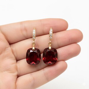 Puede incluir: Un par de pendientes dorados con grandes piedras preciosas cuadradas de color rojo intenso. Los pendientes tienen un cierre de palanca con pequeños cristales transparentes. Las piedras preciosas están facetadas, captando la luz y añadiendo brillo.