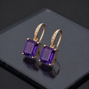 May include: A pair of gold-colored drop earrings with rectangular, deep purple gemstones. The earrings have a curved top with small, clear stones. The earrings are displayed on a reflective surface.