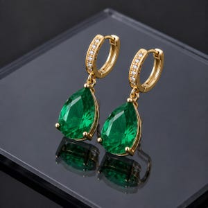 May include: A pair of gold hoop earrings featuring a hinged closure and a row of small clear stones along the front. Each earring suspends a large, faceted, pear-shaped emerald green gemstone held in a four-prong gold setting.