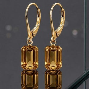May include: A pair of gold-colored leverback earrings. Each earring features a rectangular, amber-colored gemstone. The earrings are displayed against a dark background, with a reflective surface below.
