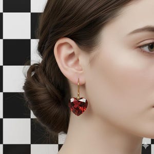 Puede incluir: Un pendiente dorado con una gema roja facetada en forma de corazón. El pendiente cuelga de un gancho simple. El fondo es un patrón de cuadros blancos y negros.