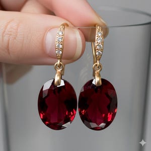 Könnte beinhalten: Ein Paar goldfarbene Ohrhänger. Jeder Ohrring hat einen großen, ovalen, facettierten, tiefroten Edelstein, der an einem goldenen Reifen mit kleinen, klaren Kristallen befestigt ist. Der Hintergrund ist unscharf.