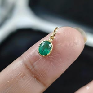 Könnte beinhalten: Ein kleiner, ovaler Smaragdanhänger mit Goldfassung. Der leuchtend grüne Edelstein ist in einen polierten Goldrahmen mit einer kleinen Schlaufe für eine Kette gefasst. Der Anhänger wird von einem Finger gehalten.