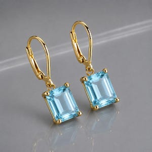 May include: A pair of gold leverback earrings, each featuring a single rectangular emerald-cut light blue gemstone suspended from a polished gold hook. The stones are clear and faceted, held in place by four small gold prongs at each corner.