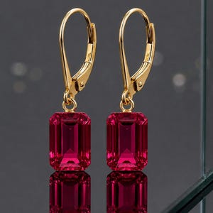 May include: A pair of gold leverback earrings featuring rectangular, deep pink gemstones. The gemstones are reflected in the surface below. The earrings have a classic, elegant design.