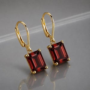 May include: A pair of gold leverback earrings featuring rectangular, deep red garnet gemstones. Each stone is held in a four-prong setting, suspended from a polished gold hook mechanism that provides a secure closure for these dangling earrings.