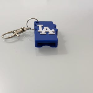 Puede incluir: Un llavero azul con un mosquet&oacute;n y un anillo plateados. El llavero tiene forma rectangular con las letras blancas "LA". El llavero est&aacute; sobre una superficie blanca.