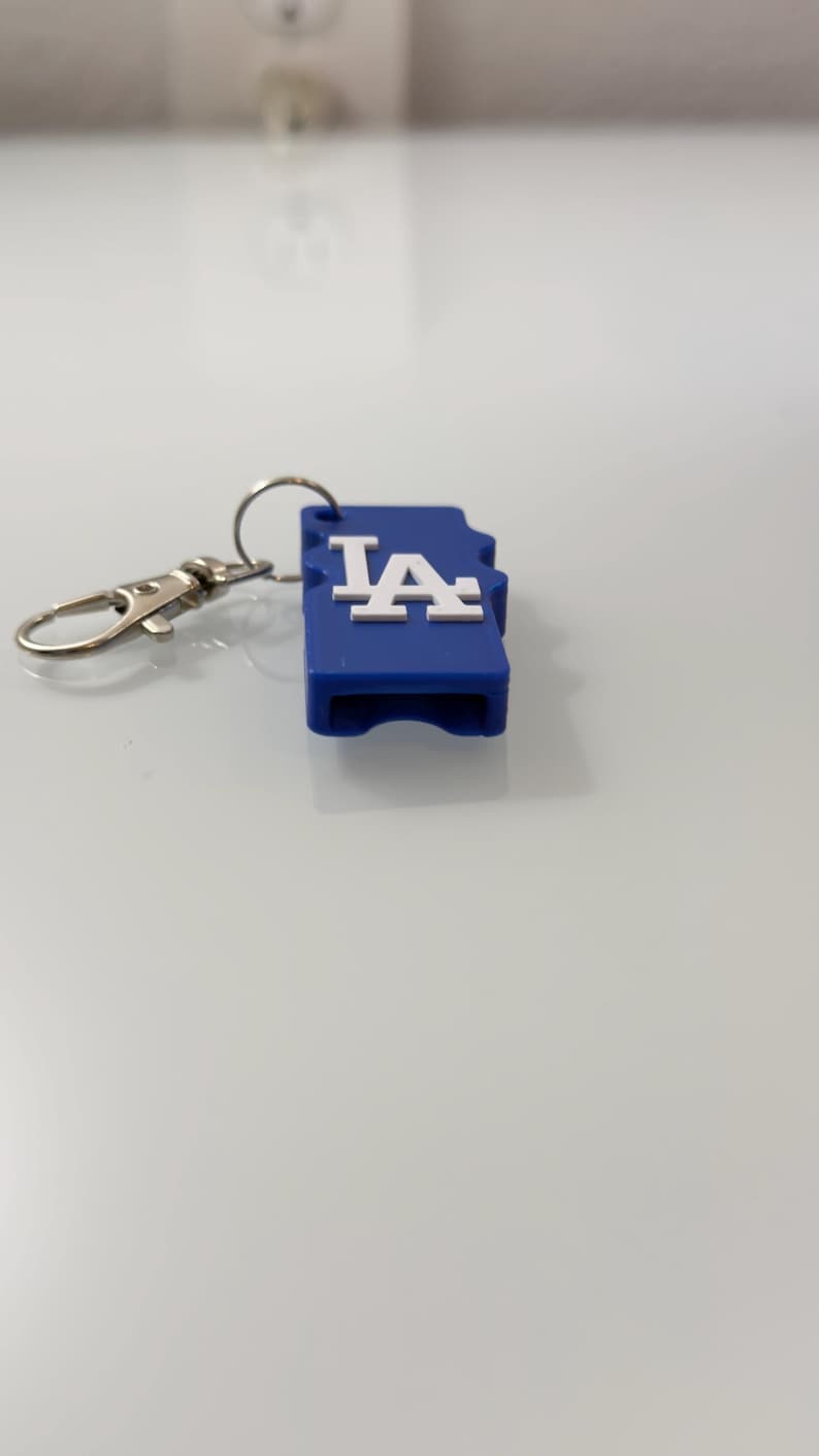 Puede incluir: Un llavero azul con un mosquet&oacute;n y un anillo plateados. El llavero tiene forma rectangular con las letras blancas "LA". El llavero est&aacute; sobre una superficie blanca.
