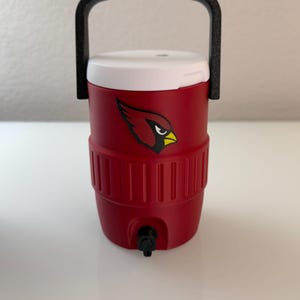 May include: Red insulated water cooler with a white lid and black handle. The cooler features a team logo of a cardinal bird. It has a black spigot for dispensing liquids.