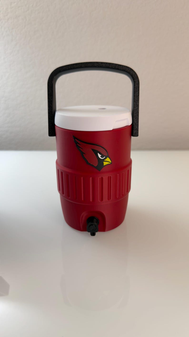 May include: Red insulated water cooler with a white lid and black handle. The cooler features a team logo of a cardinal bird. It has a black spigot for dispensing liquids.