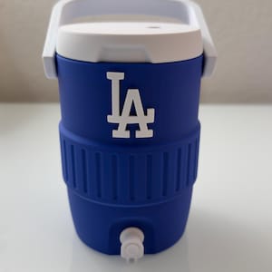 Puede incluir: Un enfriador de agua azul y blanco con el logotipo de LA. El enfriador tiene un asa y una tapa blancas, y un grifo en la parte inferior. El enfriador est&aacute; dise&ntilde;ado para mantener las bebidas fr&iacute;as.