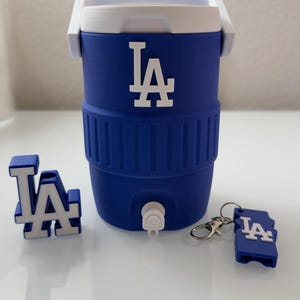 Op de afbeelding: Een blauw-witte koeler met de letters "LA" in wit. De koeler heeft een witte handgreep en een kraan. Ook inbegrepen zijn een "LA" sleutelhanger en een 3D "LA" logo.