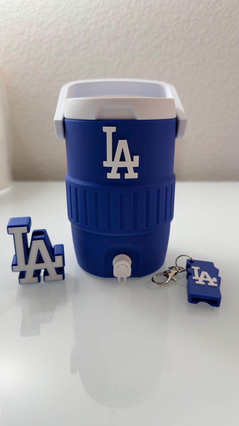 Puede incluir: Una nevera azul y blanca con las letras "LA" en blanco. La nevera tiene un asa blanca y un grifo. Tambi&eacute;n se incluyen un llavero "LA" y un logotipo "LA" en 3D.
