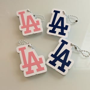 May include: Four white keychains featuring the Los Angeles Dodgers LA logo. Two keychains have a pink logo and two have a dark blue logo. Each keychain is attached to a small silver-toned metal ball chain for easy attachment to keys or bags.