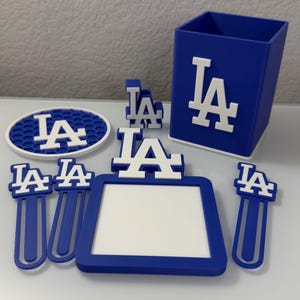 May include: A collection of blue and white Los Angeles Dodgers themed accessories. Includes a pen holder, picture frame, bookmarks, and a coaster, all featuring the team's logo.