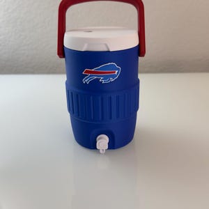 May include: A blue and white water cooler with a red handle. The cooler features the Buffalo Bills logo on the front. It has a white lid and a spigot for dispensing liquids. The cooler is designed to keep drinks cold.