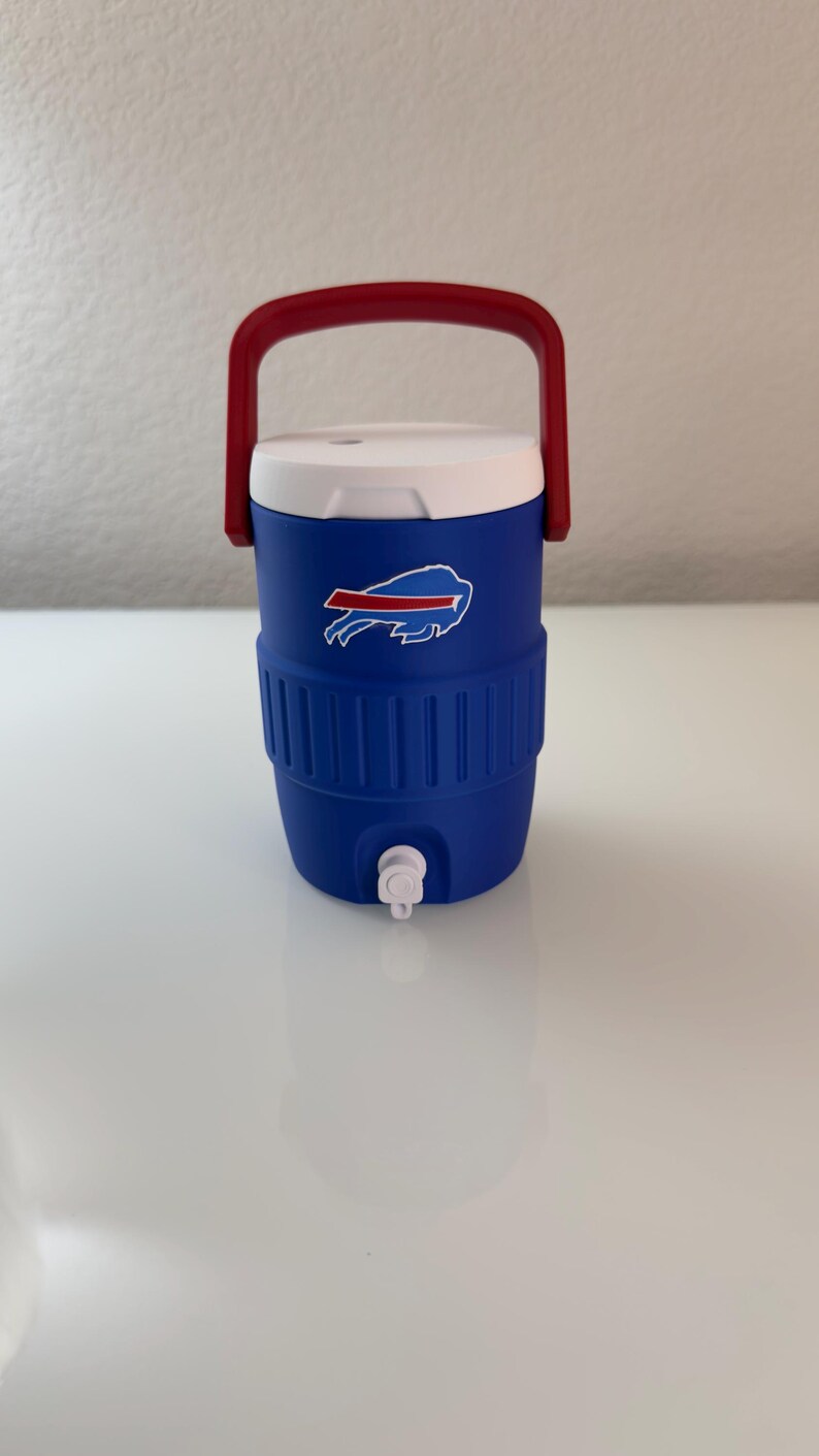 May include: A blue and white water cooler with a red handle. The cooler features the Buffalo Bills logo on the front. It has a white lid and a spigot for dispensing liquids. The cooler is designed to keep drinks cold.