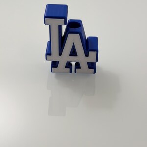 Puede incluir: Logotipo "LA" azul y blanco, probablemente del equipo de b&eacute;isbol Los Angeles Dodgers. Las letras son blancas con contorno azul y fondo azul. El objeto es probablemente un art&iacute;culo decorativo o un peque&ntilde;o accesorio.