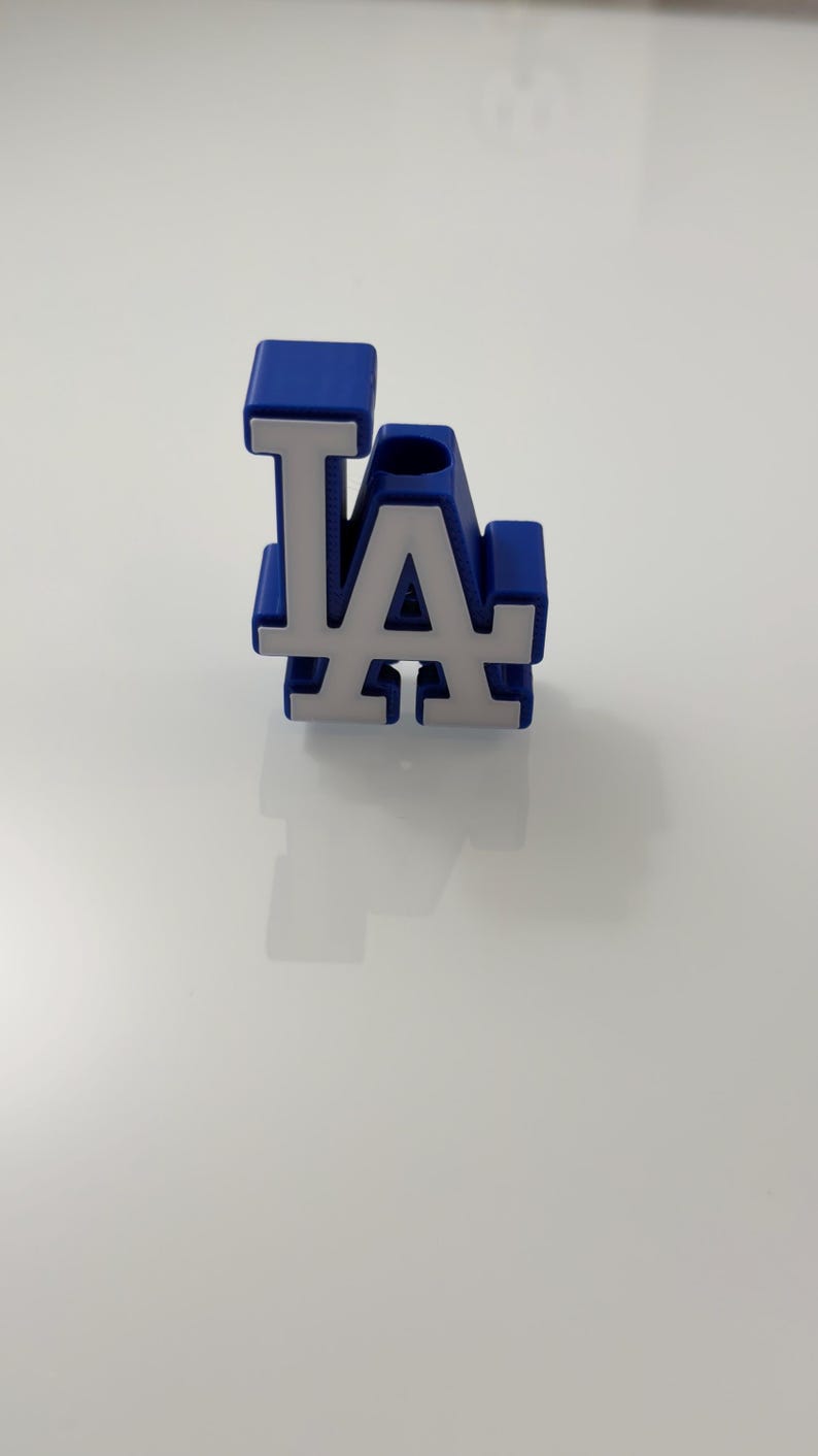 Puede incluir: Logotipo "LA" azul y blanco, probablemente del equipo de b&eacute;isbol Los Angeles Dodgers. Las letras son blancas con contorno azul y fondo azul. El objeto es probablemente un art&iacute;culo decorativo o un peque&ntilde;o accesorio.