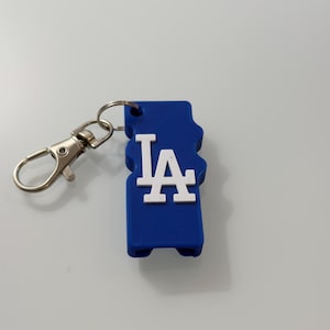 Puede incluir: Un llavero azul con las letras blancas "LA" en una fuente estilizada. El llavero tiene un cierre y un anillo de color plateado para sujetarlo a llaves o a un bolso. El dise&ntilde;o es un logotipo de un equipo deportivo.