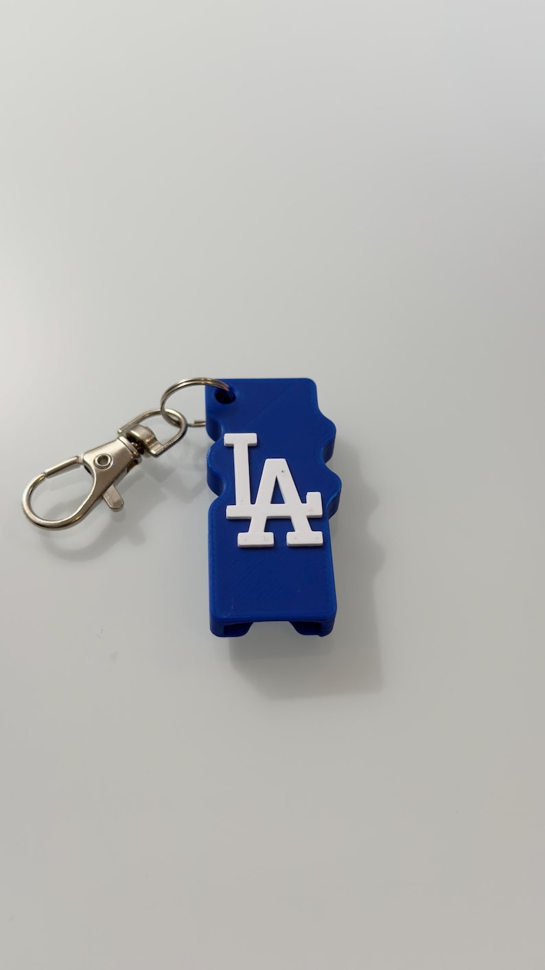 Puede incluir: Un llavero azul con las letras blancas "LA" en una fuente estilizada. El llavero tiene un cierre y un anillo de color plateado para sujetarlo a llaves o a un bolso. El dise&ntilde;o es un logotipo de un equipo deportivo.