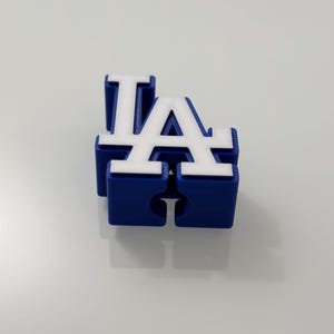 Puede incluir: Un logotipo "LA" azul y blanco impreso en 3D, probablemente para el equipo de b&eacute;isbol de los Dodgers de Los &Aacute;ngeles. Las letras son de estilo bloque, con letras blancas sobre una base azul. El objeto est&aacute; sobre una superficie blanca.