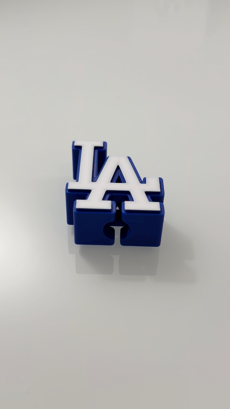 Puede incluir: Un logotipo "LA" azul y blanco impreso en 3D, probablemente para el equipo de b&eacute;isbol de los Dodgers de Los &Aacute;ngeles. Las letras son de estilo bloque, con letras blancas sobre una base azul. El objeto est&aacute; sobre una superficie blanca.