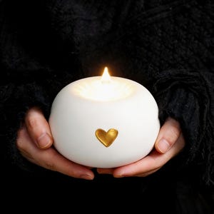 Puede incluir: Un portavelas esférico blanco con un emblema de corazón dorado. El portavelas emite un brillo cálido y suave de un LED en forma de llama. El diseño es simple y elegante, adecuado para la decoración del hogar.
