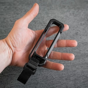 May include: A black rectangular phone case with a strap is held in a hand. The case has a geometric design and a textured strap. The background is a gray surface.