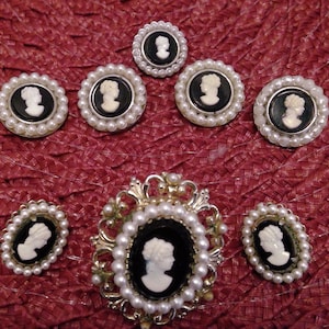 May include: A set of eight cameo brooches with white cameo portraits on black backgrounds. The brooches are surrounded by white pearls and are arranged in a circular pattern. The brooches are on a red background.