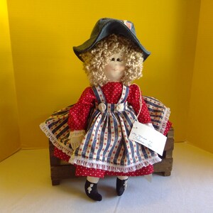 May include: A handmade rag doll wearing a red and blue striped dress with white stars, a blue denim hat, and black shoes. The doll is sitting on a wooden crate with a tag that reads "Grandma's Creations".