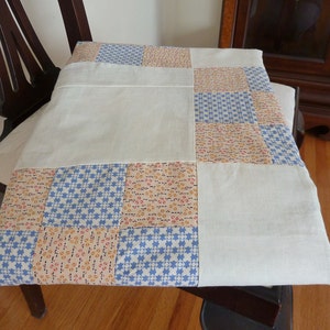 May include: A patchwork quilt with a white background and squares of blue, orange, and white fabric. The quilt is folded and placed on a wooden chair.