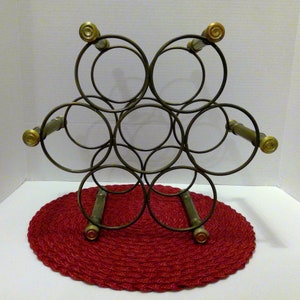 May include: A black metal wine rack with six circular openings and gold-colored accents. The rack is sitting on a red woven mat.