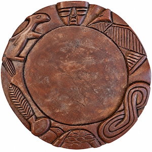 Puede incluir: Una placa circular de piedra marrón con un disco central liso rodeado por un borde tallado. Los relieves incluyen un rostro humano, un ave, un patrón geométrico, una forma de hoja, un diseño en espiral y una figura de tortuga.