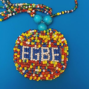 Puede incluir: Un collar de cuentas con un colgante circular. El colgante presenta la palabra "EGBE" en cuentas azules sobre un fondo de cuentas rojas, amarillas y blancas. El collar tiene un fondo azul y una cadena de cuentas con cuentas turquesas.