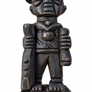 Puede incluir: Una figura de madera tallada a mano de color marrón oscuro que representa a un personaje con gorra y pañuelo al cuello. La figura se apoya sobre una base rectangular, sosteniendo un objeto cilíndrico largo en la mano derecha y un objeto más pequeño y redondeado en la mano izquierda.