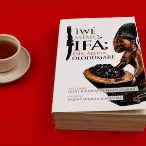 Puede incluir: Un libro titulado "Ìwé Mímọ́ Ifá: Ẹ̀sìn Àkọ́dá Olódùmarè" con una cubierta blanca que presenta la ilustración de una persona sosteniendo un cuenco con objetos oscuros, junto a una taza de té de cerámica clara con líquido marrón oscuro.