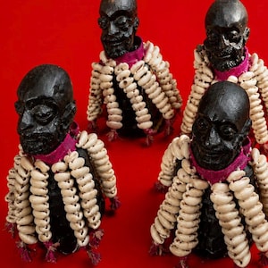 Puede incluir: Cuatro bustos de madera tallada de color marrón oscuro con rasgos faciales humanos. Cada busto está adornado con un cuello de tela magenta vibrante, decorado con múltiples hilos de conchas de cauri color crema en forma de flecos.
