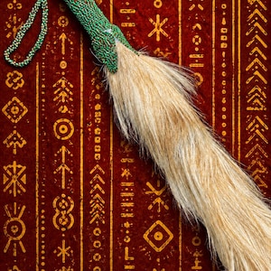 Pode incluir: Um espanta-moscas com um cabo coberto por pequenas contas verde-vibrantes e um longo tufo fluido de pelo animal castanho-claro. O cabo inclui uma alça de contas a condizer sobre um fundo vermelho profundo com símbolos dourados.