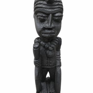 Puede incluir: Una figura de madera tallada a mano con un acabado gris oscuro. La escultura representa a un personaje de pie con una cabeza prominente, rasgos faciales detallados y un adorno texturizado en el cuello. Sostiene un objeto tipo bastón en la mano.