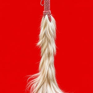 Pode incluir: Um espanta-moscas com um cabo coberto por intrincados trabalhos em missangas vermelhas e brancas, com uma pequena argola de missangas presa no topo. O cabo está ligado a um longo e fluido feixe de fibras de pelo animal loiro claro.