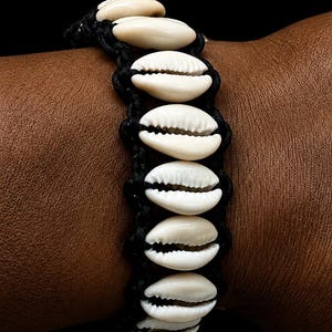 Puede incluir: Una pulsera que presenta una serie de conchas de cauri de color blanco roto dispuestas en una fila vertical, aseguradas por un cordón negro anudado. Las conchas son lisas y de forma ovalada con una ranura central, creando un contraste.