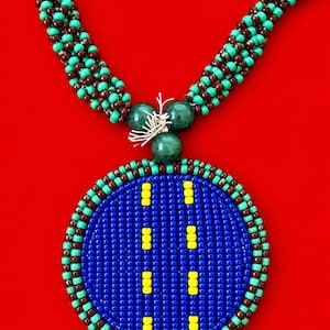 Puede incluir: Un collar de cuentas con un gran colgante circular de fondo azul, dos filas verticales de cuentas amarillas y un borde de cuentas granates y turquesas. La cadena del collar consta de varios hilos de cuentas granates y turquesas.